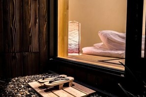 Una habitación con piso de madera, cama con ropa blanca, ventana con vista a un edificio y una pequeña mesa de madera con zapatillas.