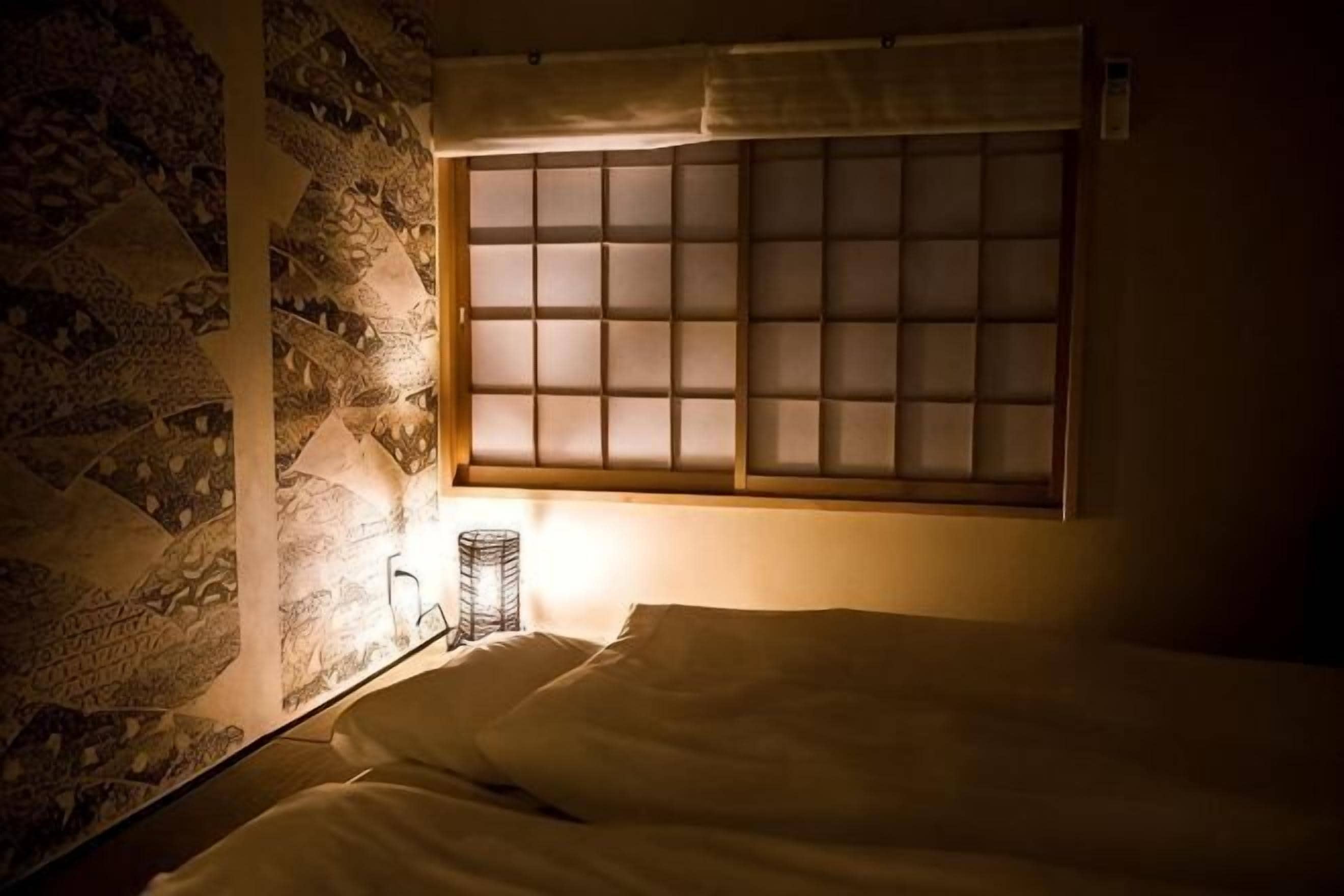 A room with a bed, a window with a grid pattern, a wall with a geometric pattern, and a small table with a lamp.