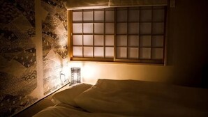 A room with a bed, a window with a grid pattern, a wall with a geometric pattern, and a small table with a lamp.