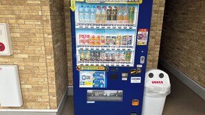 Een automaat met verschillende drankjes en snacks, een touchscreen en een prullenbak.