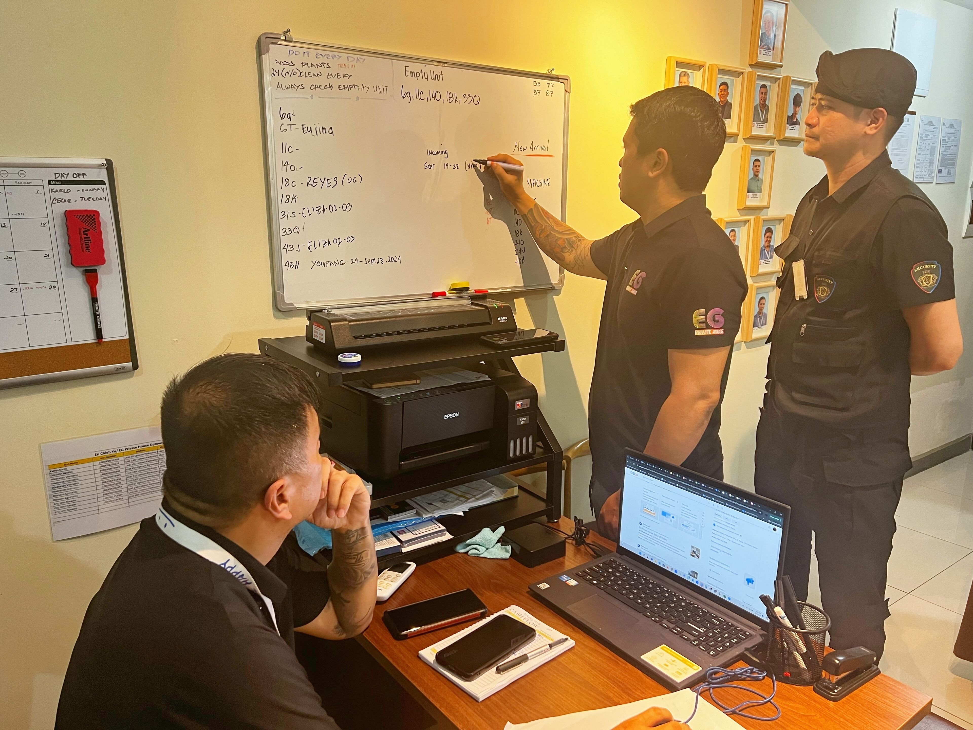 A security officer writing on a whiteboard in a room with a desk, laptop, and various office supplies.