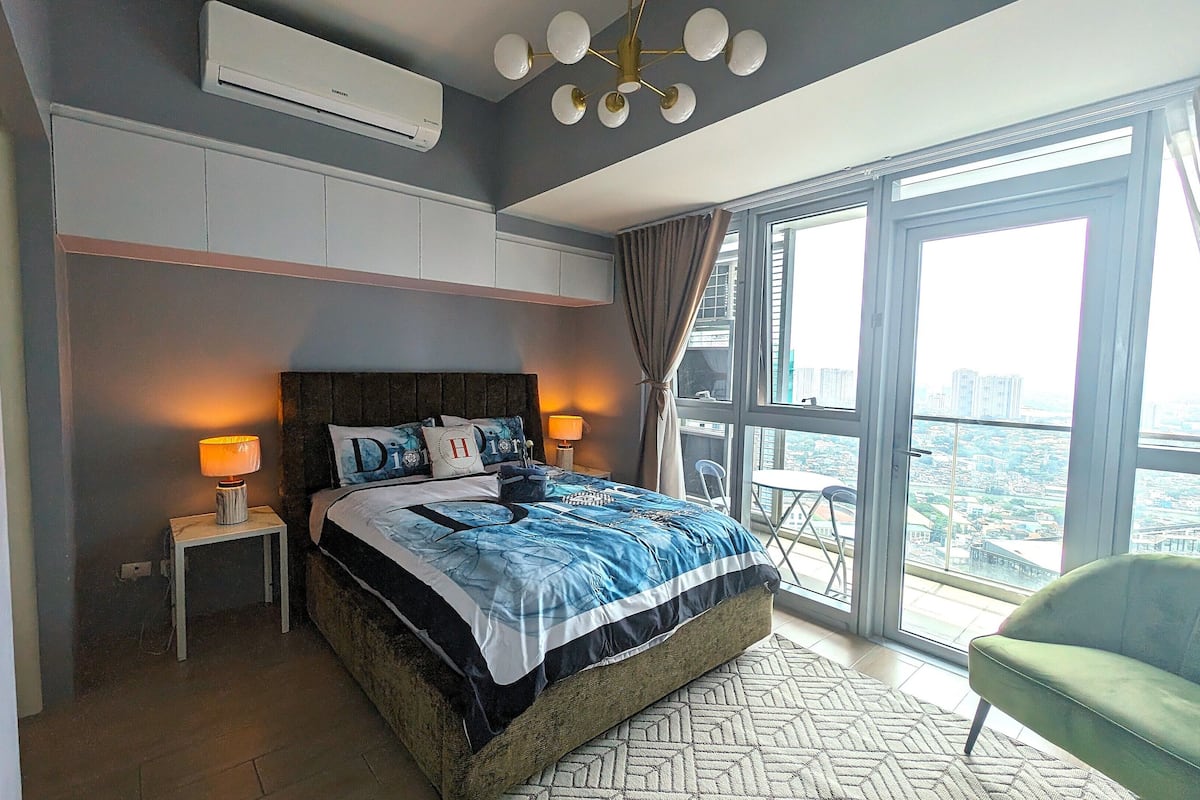 A modern bedroom with a large bed, bedside lamps, a view of the city through large windows, and a balcony chair.