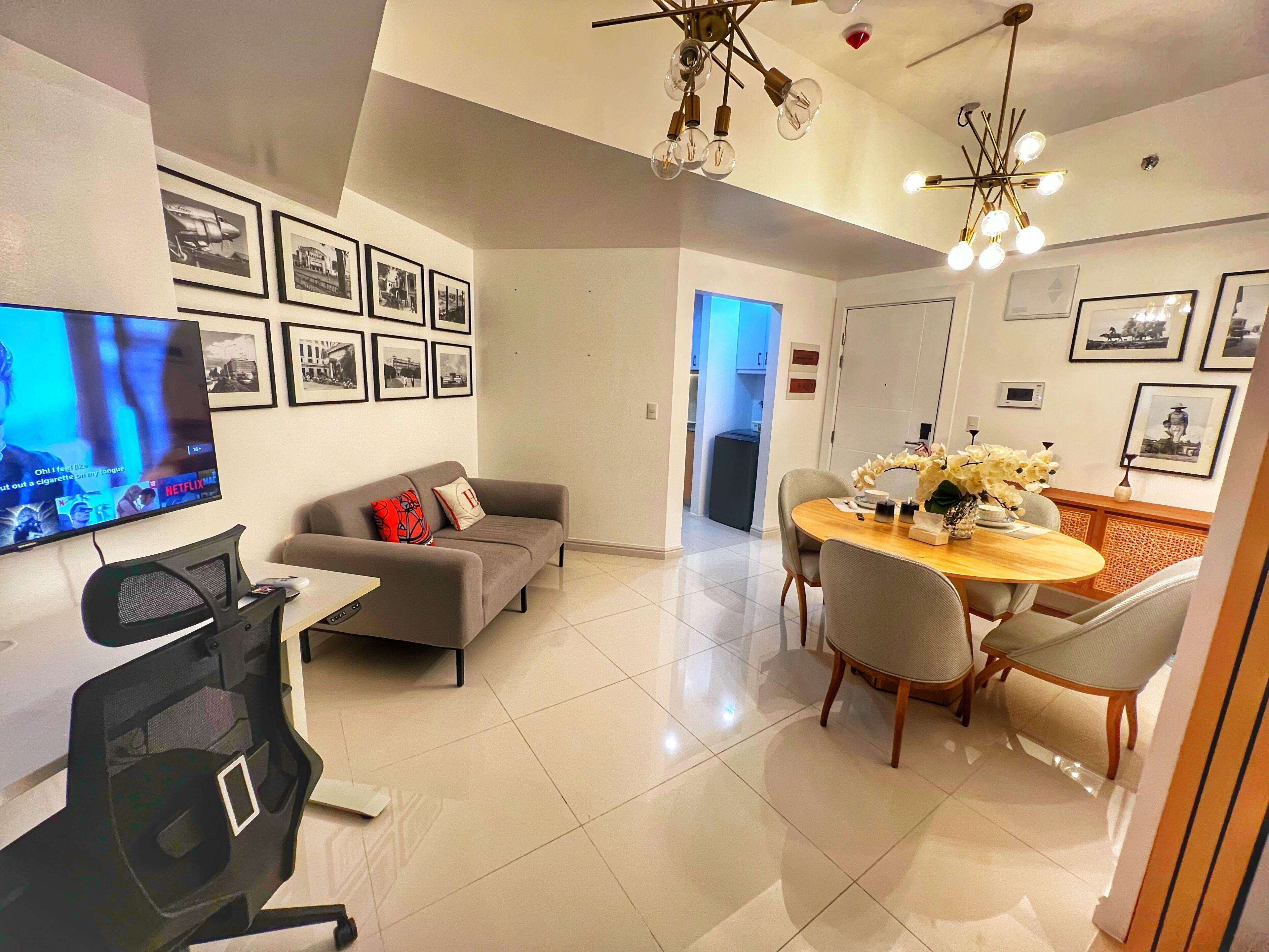 Una sala moderna con televisor de pantalla plana, un sofá gris, una mesa de comedor con sillas y una cocina al fondo.