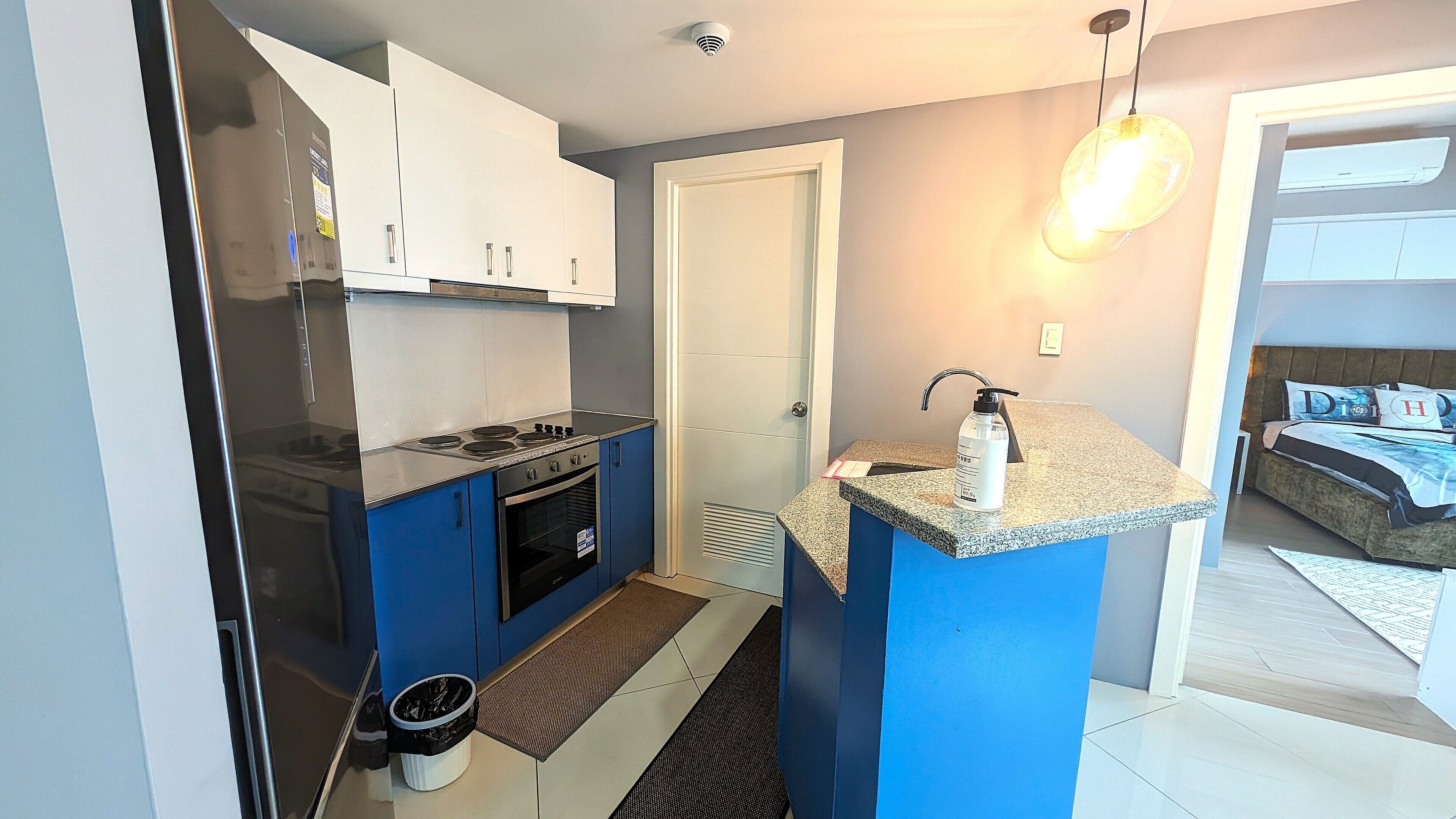 A compact kitchen with a stove, oven, and blue cabinets. There is a small trash bin and a countertop with a soap dispenser. A door leads to a bedroom with a bed and a nightstand.