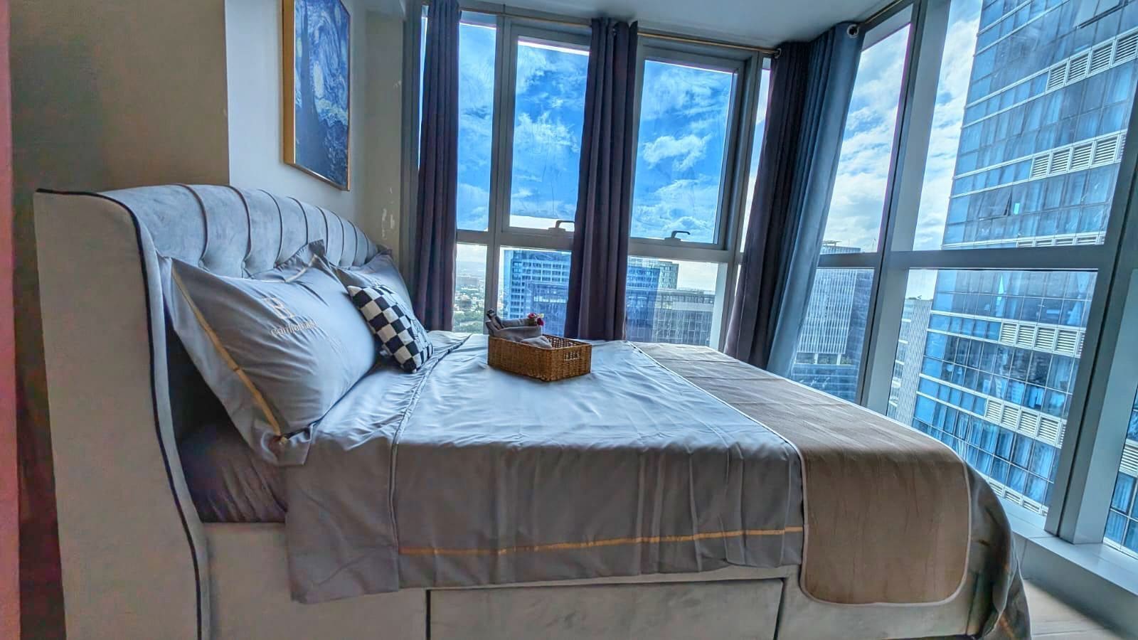 A bedroom with a large bed, a window with curtains, and a view of a cityscape.