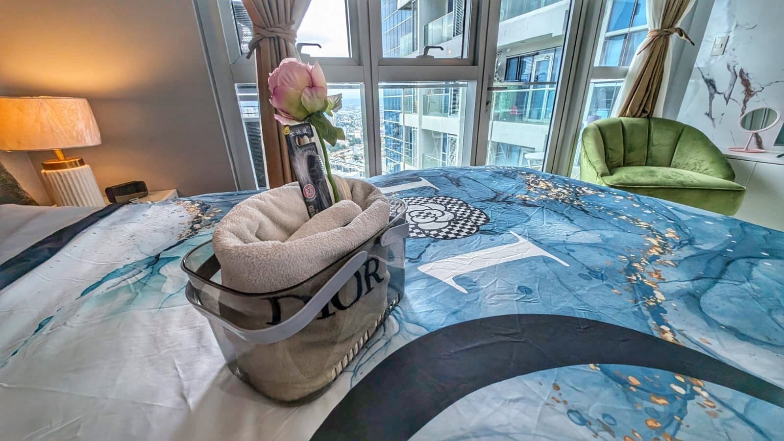 A hotel room with a bed, a green chair, a window with a view of buildings, and a lamp on a bedside table.
