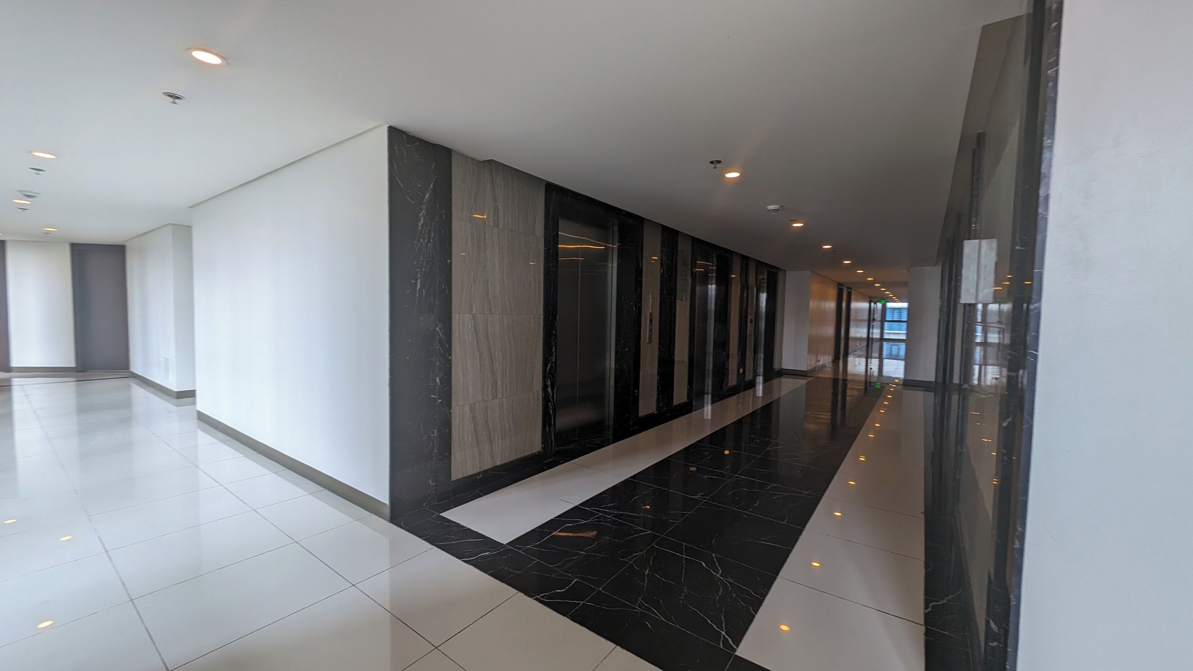 A modern hallway with a glossy black and white marble floor, recessed lighting, and a glass elevator.