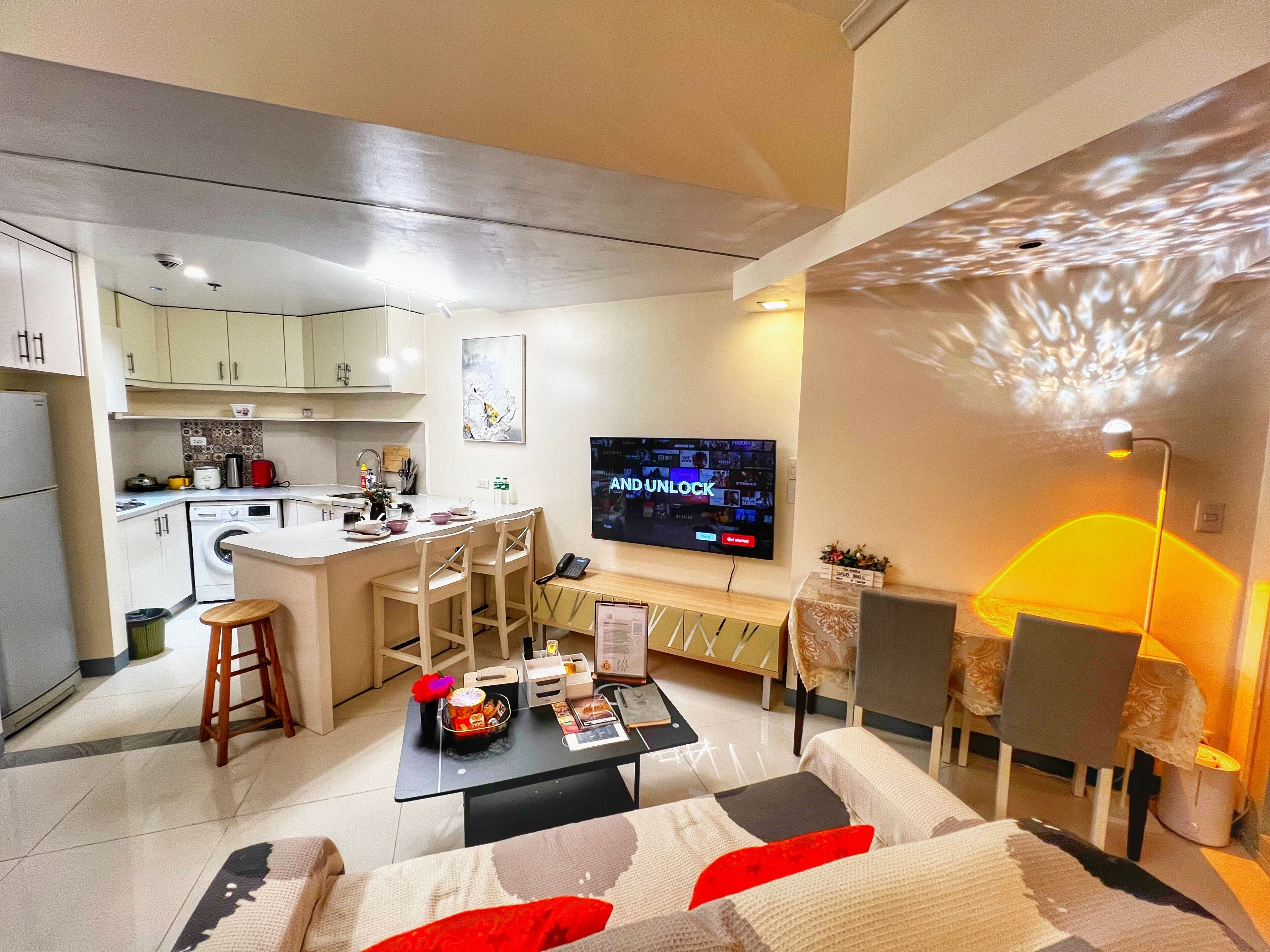 A modern living room with a sofa, coffee table, and TV. There is a kitchen area with cabinets and appliances.