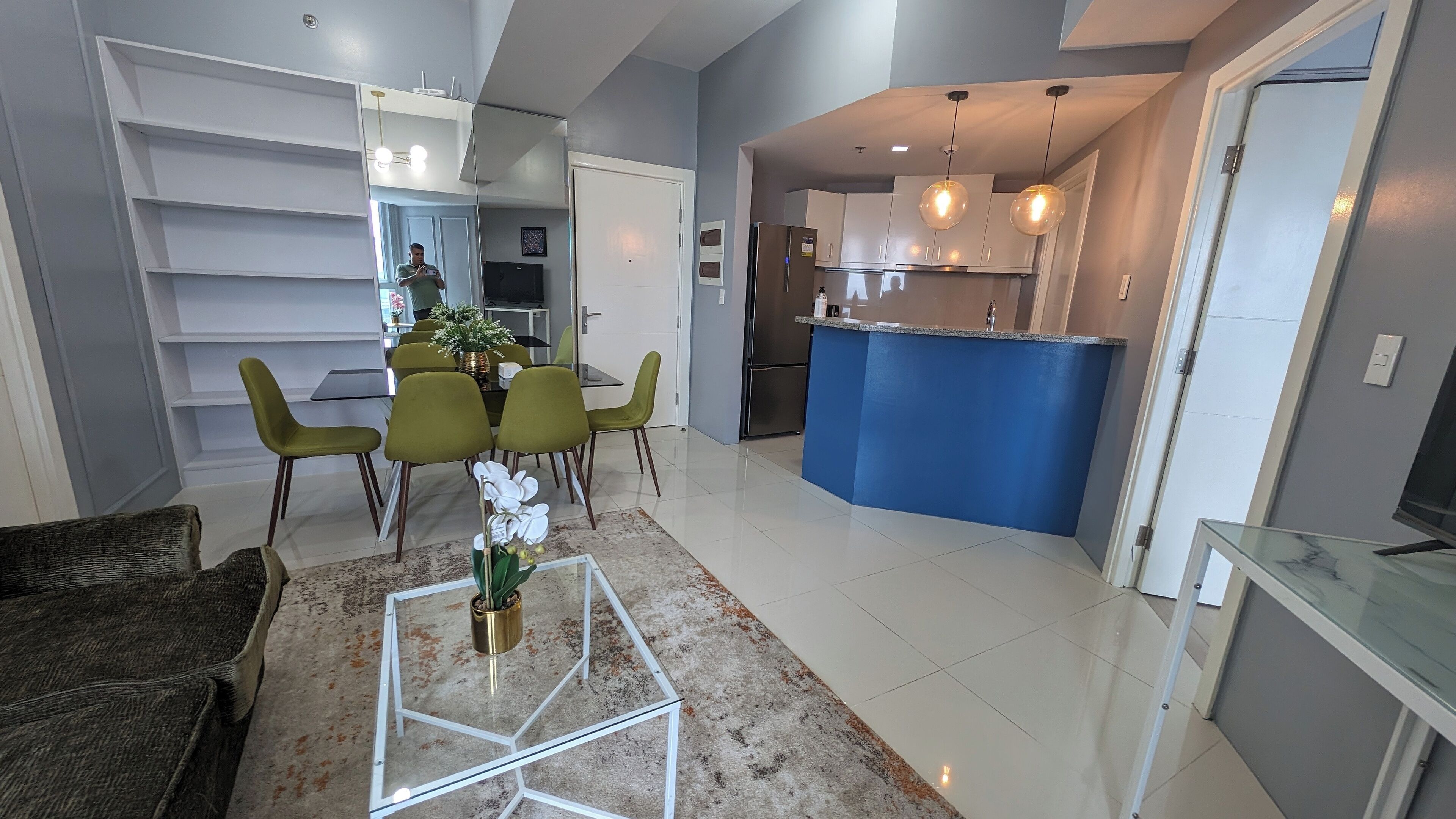 A modern living room with a glass coffee table, a dining area with green chairs, and a kitchen with a blue accent wall and stainless steel appliances.