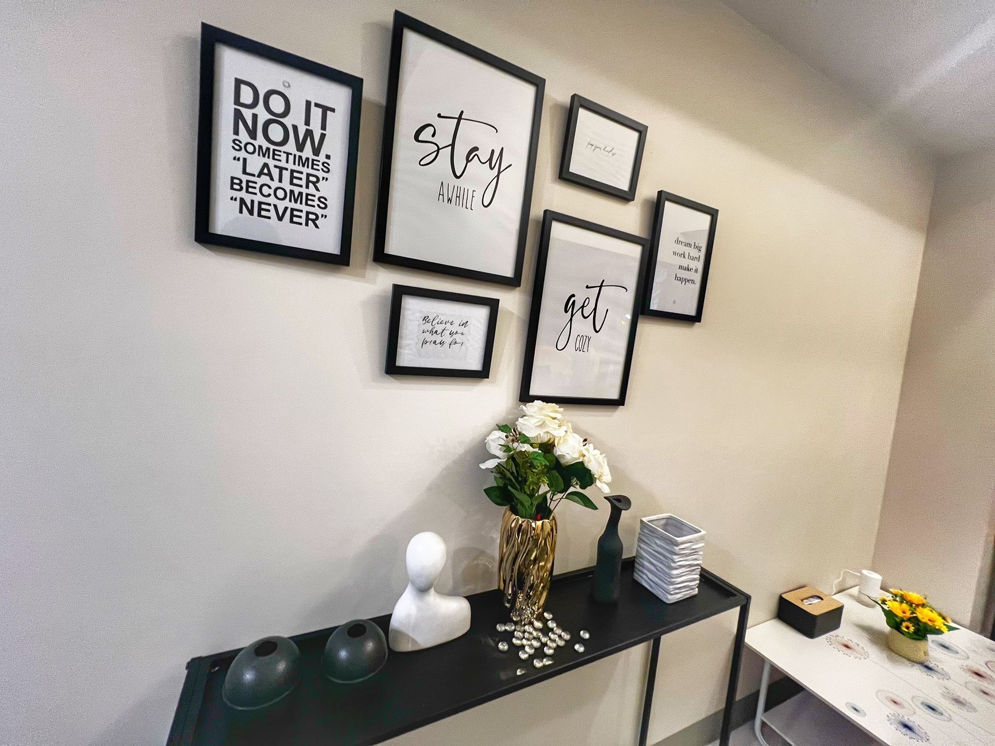 Una parete decorata con citazioni motivazionali in cornici, un vaso bianco con fiori e un consolle nero con oggetti decorativi.