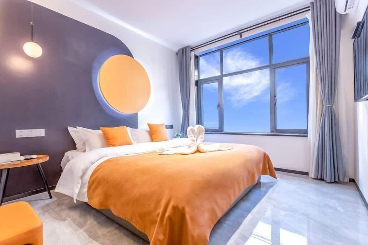 A modern hotel room with a large bed, a round mirror, a small table, and a view of the sky through a large window.
