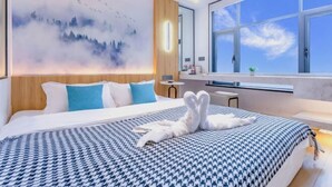 Une chambre d’hôtel moderne avec un grand lit, une vue sur le ciel par une grande fenêtre et une œuvre d’art fixée au mur représentant un paysage montagneux.
