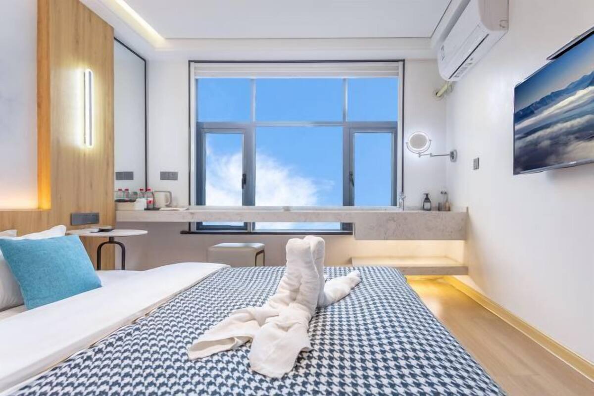 A modern hotel room with a large window, a bed with a patterned bedspread, a small table, and a mirror on the wall.