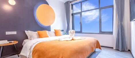 A modern hotel room with a large bed, a round wall decoration, a view of the sky through a large window, and a small table with a chair.
