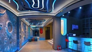 Un hall d’hôtel moderne doté d’un plafond au design original, d’un bar avec des tabourets et d’un grand écran mural.