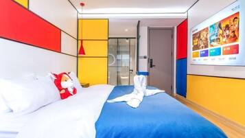 A hotel room with a bed, a wall-mounted TV displaying movie posters, and a glass door leading to a bathroom.