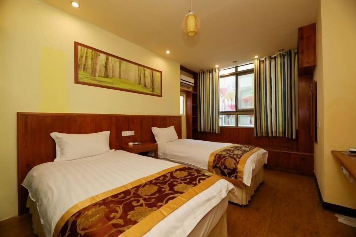 A hotel room with two beds, wooden headboards, a painting on the wall, a window with curtains, and a small desk with a chair.