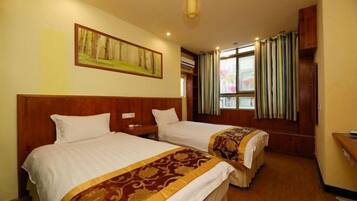 A hotel room with two beds, wooden headboards, a painting on the wall, a window with curtains, and a small desk with a chair.