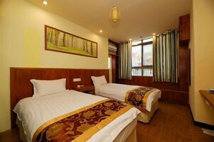 Habitación de hotel con dos camas, cabeceros de madera, un cuadro en la pared, una ventana con cortinas y un escritorio pequeño con una silla.