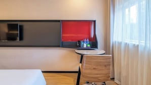 Een hotelkamer met een bed, een tafeltje met een stoel, een rode accentmuur, een televisie en een raam met gordijnen.