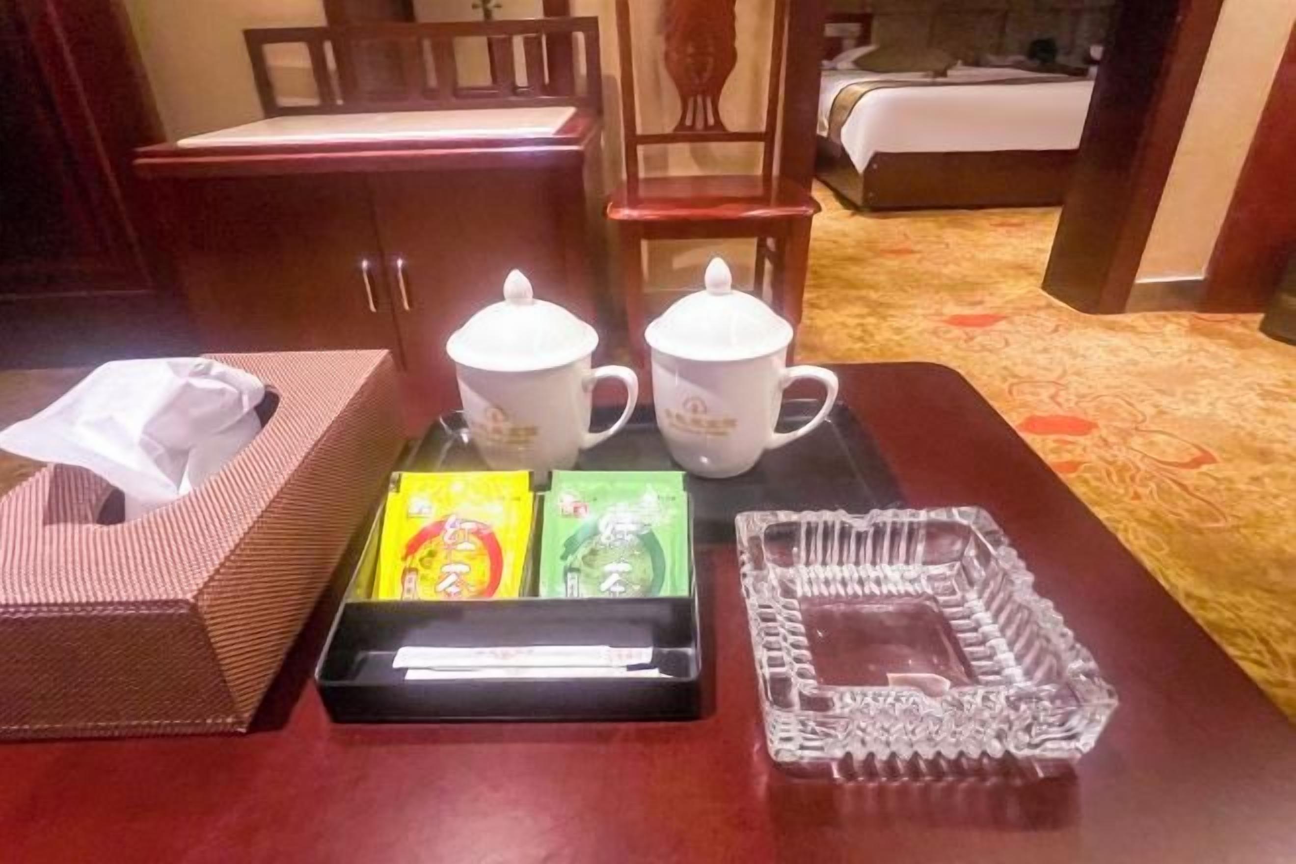 A hotel room with a wooden table, two white mugs, a tissue box, and a tray with items.