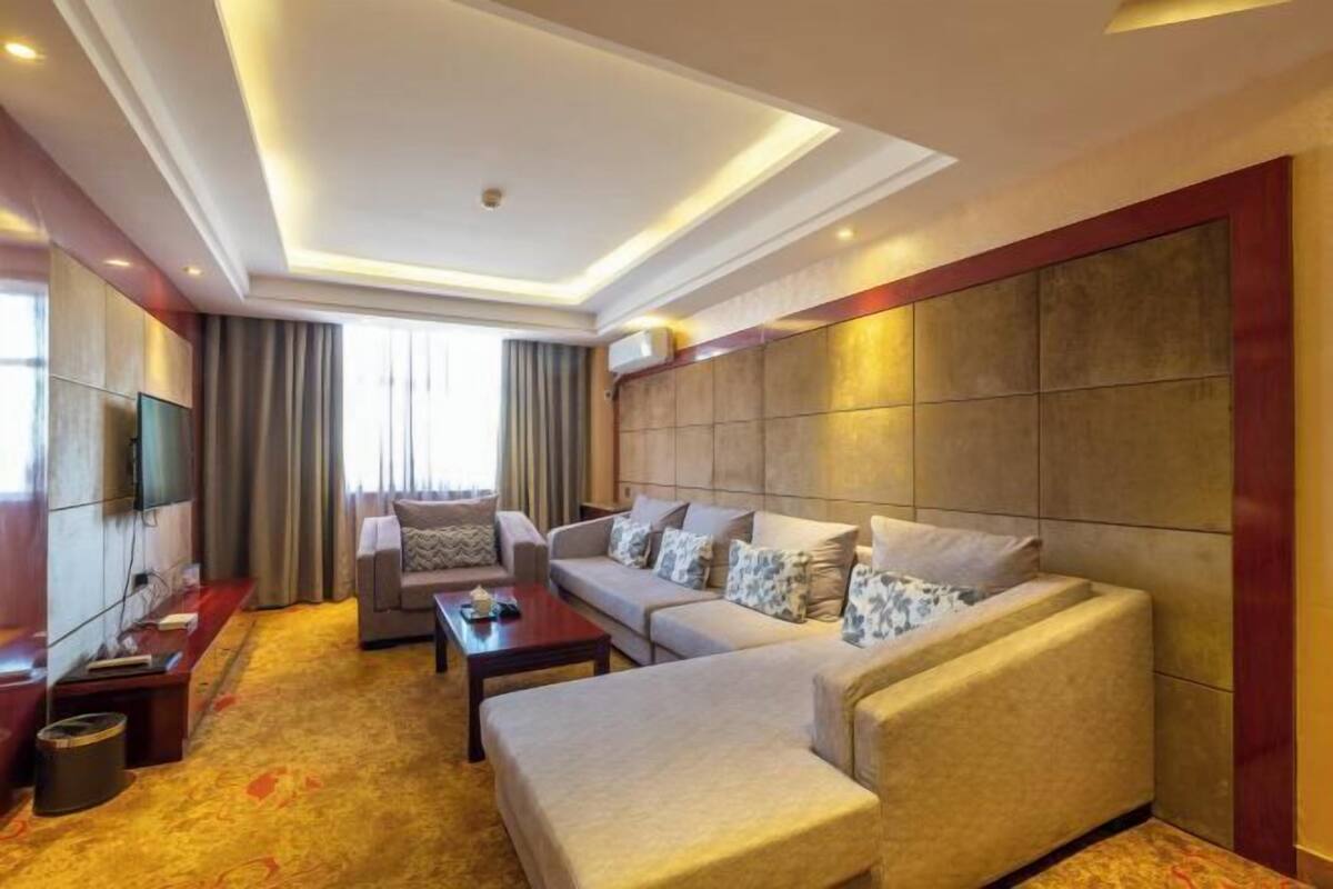 A modern hotel room with a large sofa set, a flat-screen TV, and a large window with curtains.