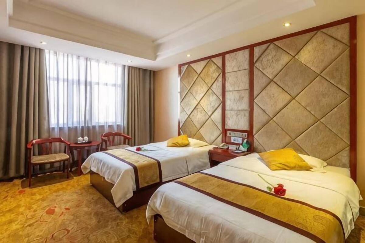 A hotel room with two beds, a small table, two chairs, and a large window with curtains.