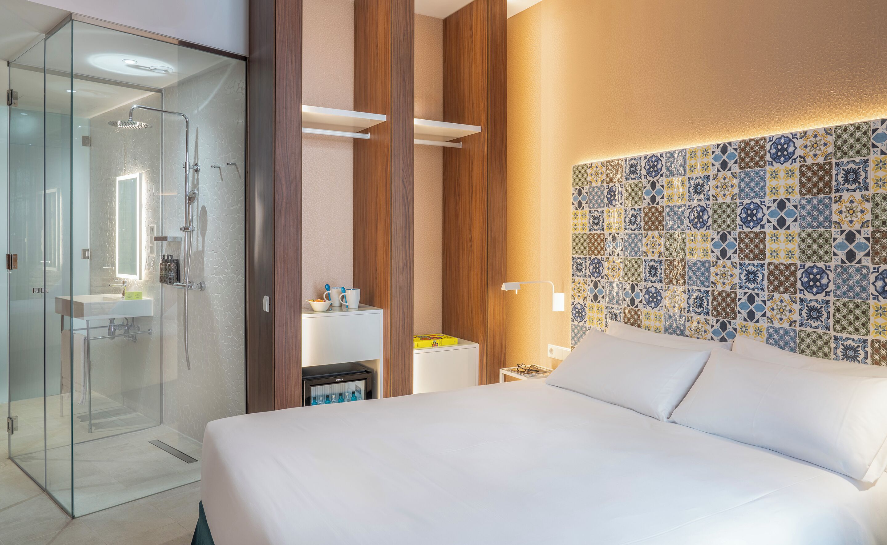 Une chambre d’hôtel dotée d’une douche en verre, d’un lit avec du linge de maison blanc, d’une table de chevet avec une lampe et d’un mur décoratif recouvert de carreaux.