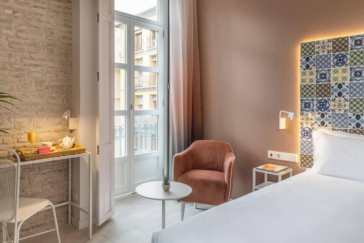 A hotel room with a brick wall, a large window, a bed, a chair, a small table with a plant, and a decorative wall.