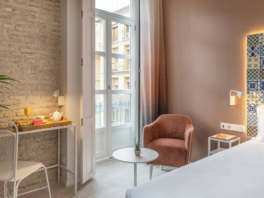 Habitación de hotel con pared de ladrillo, un ventanal grande, cama, una silla, una mesita con planta y decoración en la pared.