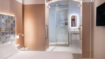 Una habitación de hotel moderna con una cama grande, zona de ducha con paredes de vidrio, una mesita de noche pequeña con una planta y un revoque de azulejos.