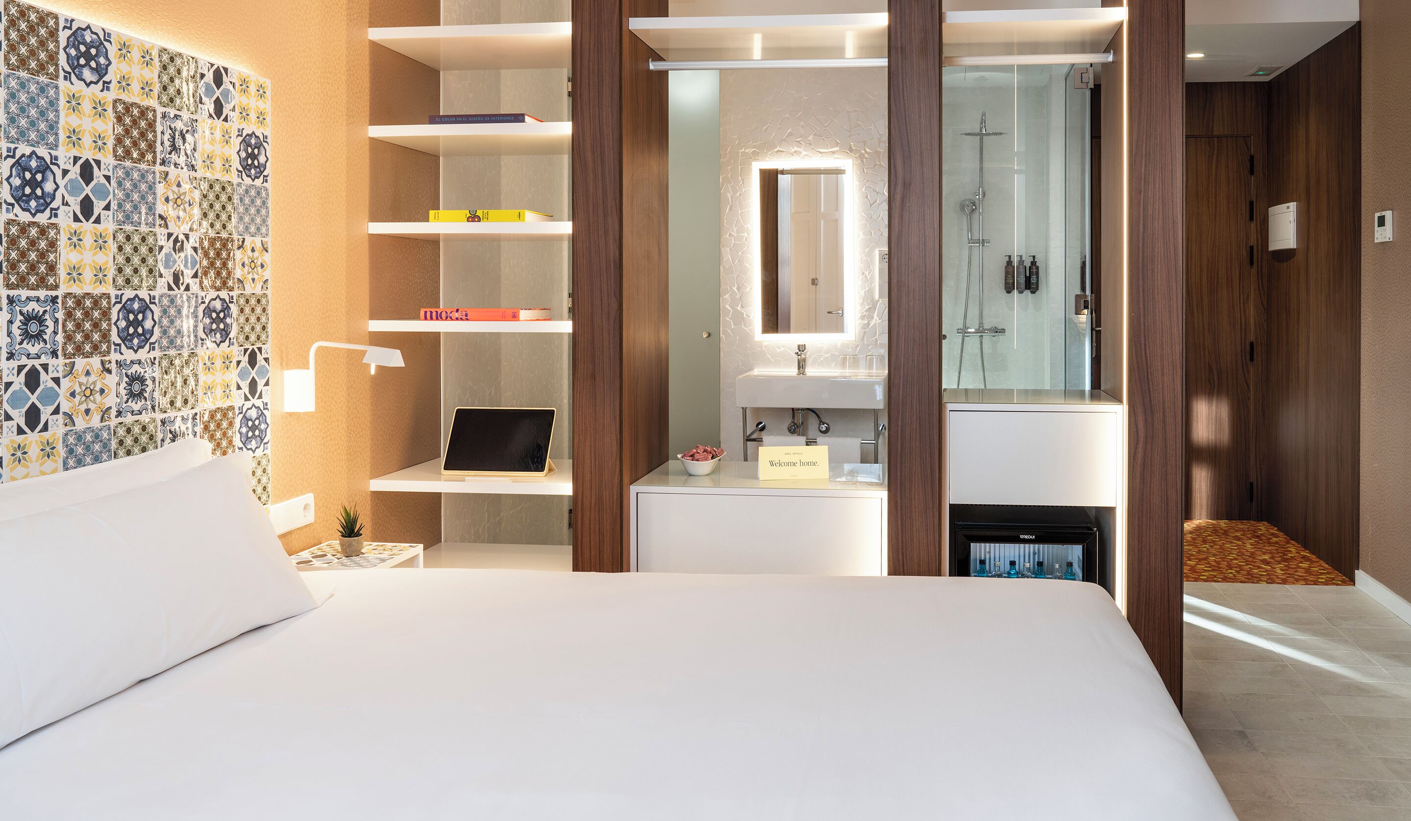 Un hotel moderno con cama, escritorio con ordenador portátil, planta pequeña, frutero, tocador con espejo y zona de ducha.