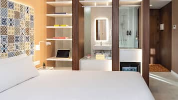 Un hotel moderno con cama, escritorio con ordenador portátil, planta pequeña, frutero, tocador con espejo y zona de ducha.