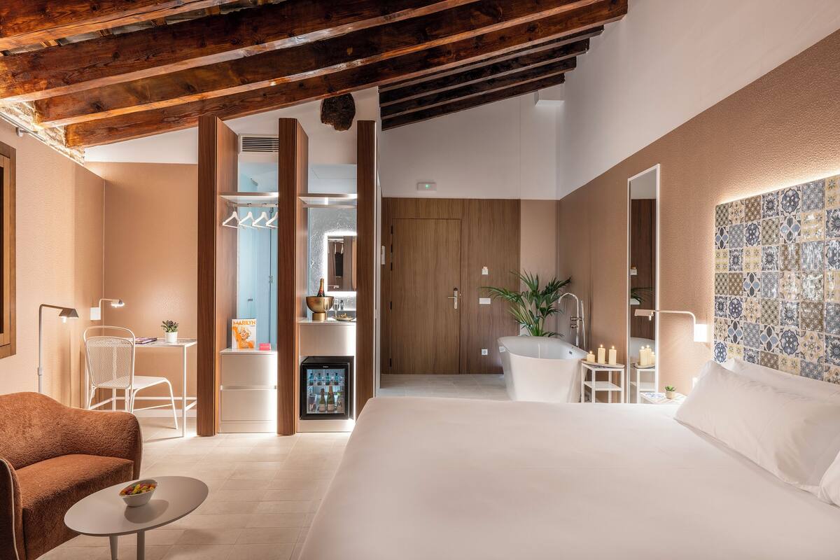 A modern hotel room with a large bed, a seating area with a small table, a wardrobe, and a bathroom with a mirror and sink.