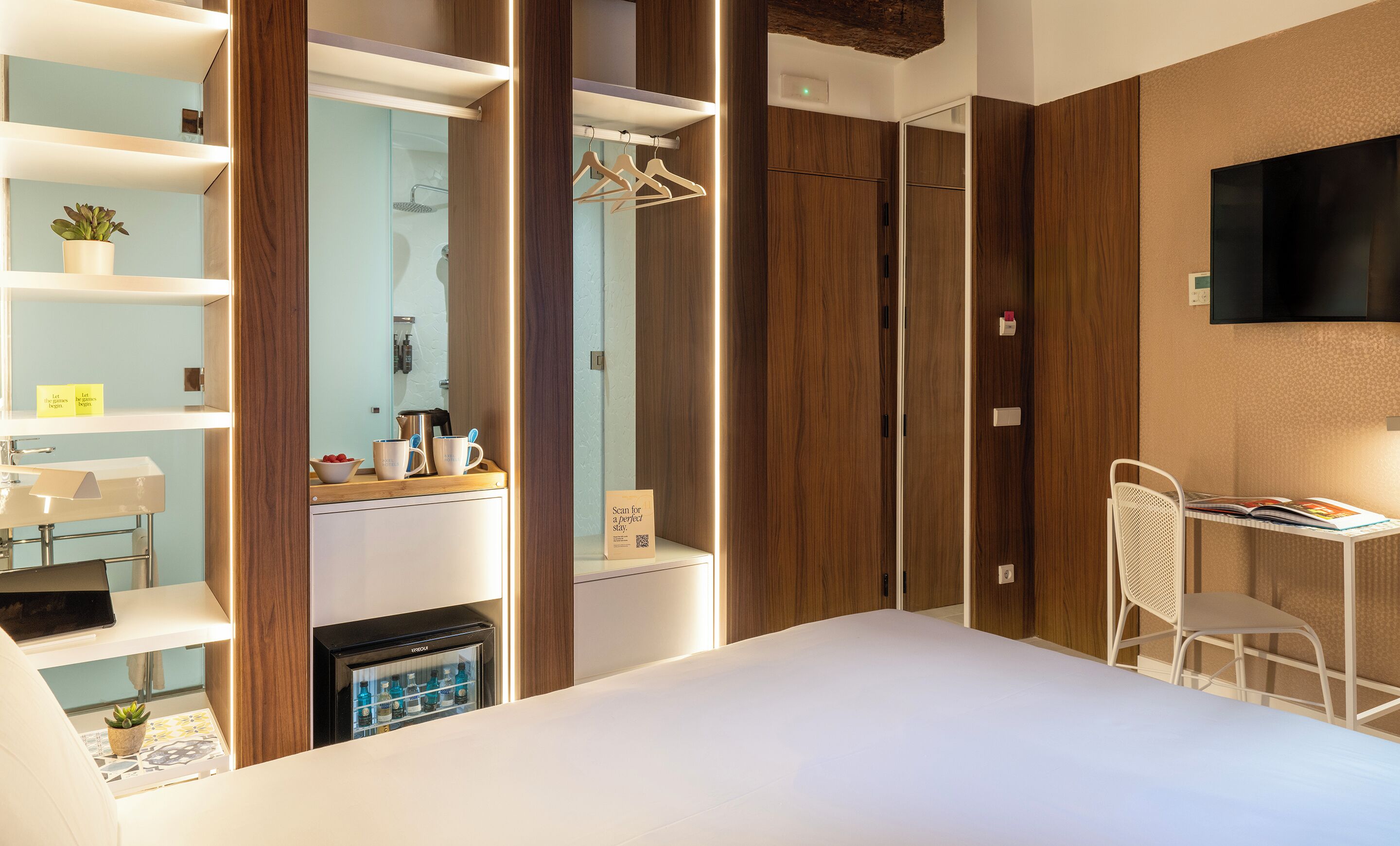 A modern hotel room with a bed, a desk with a chair, a minibar, a shower area, and wooden paneling.