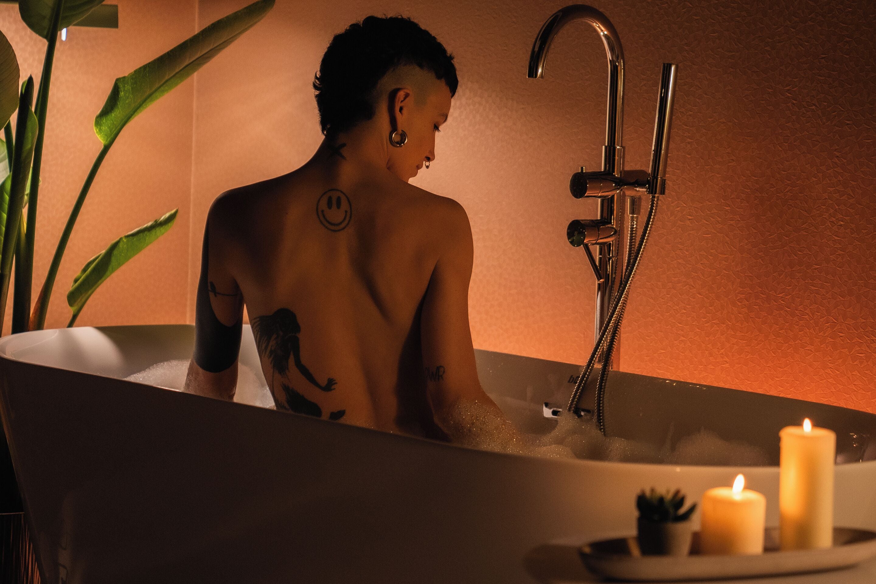 A person in a bathtub with a smiley face tattoo, surrounded by plants and candles.