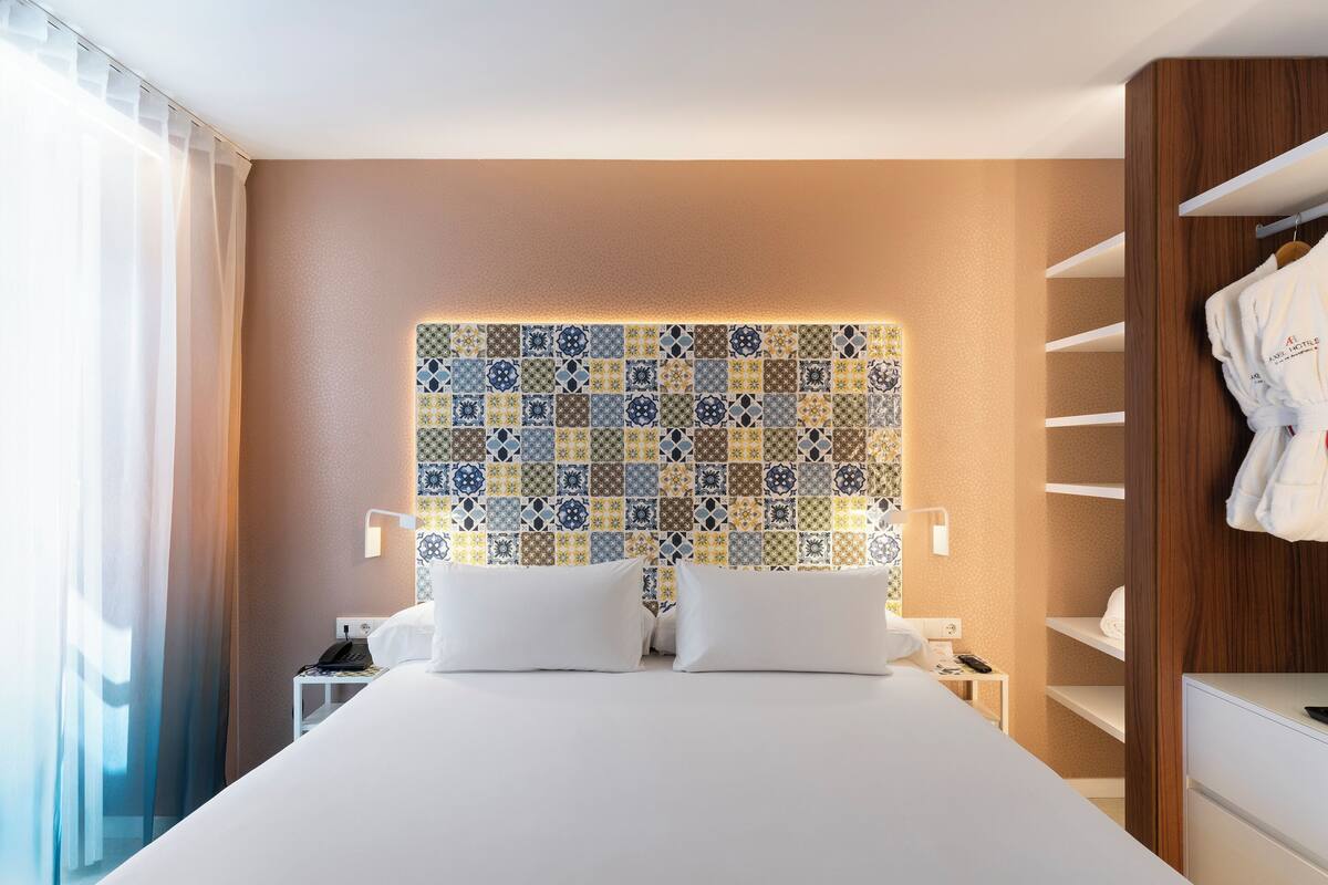 A hotel room with a large bed, a patterned headboard, a bedside table with a lamp, and a wardrobe with hanging robes.