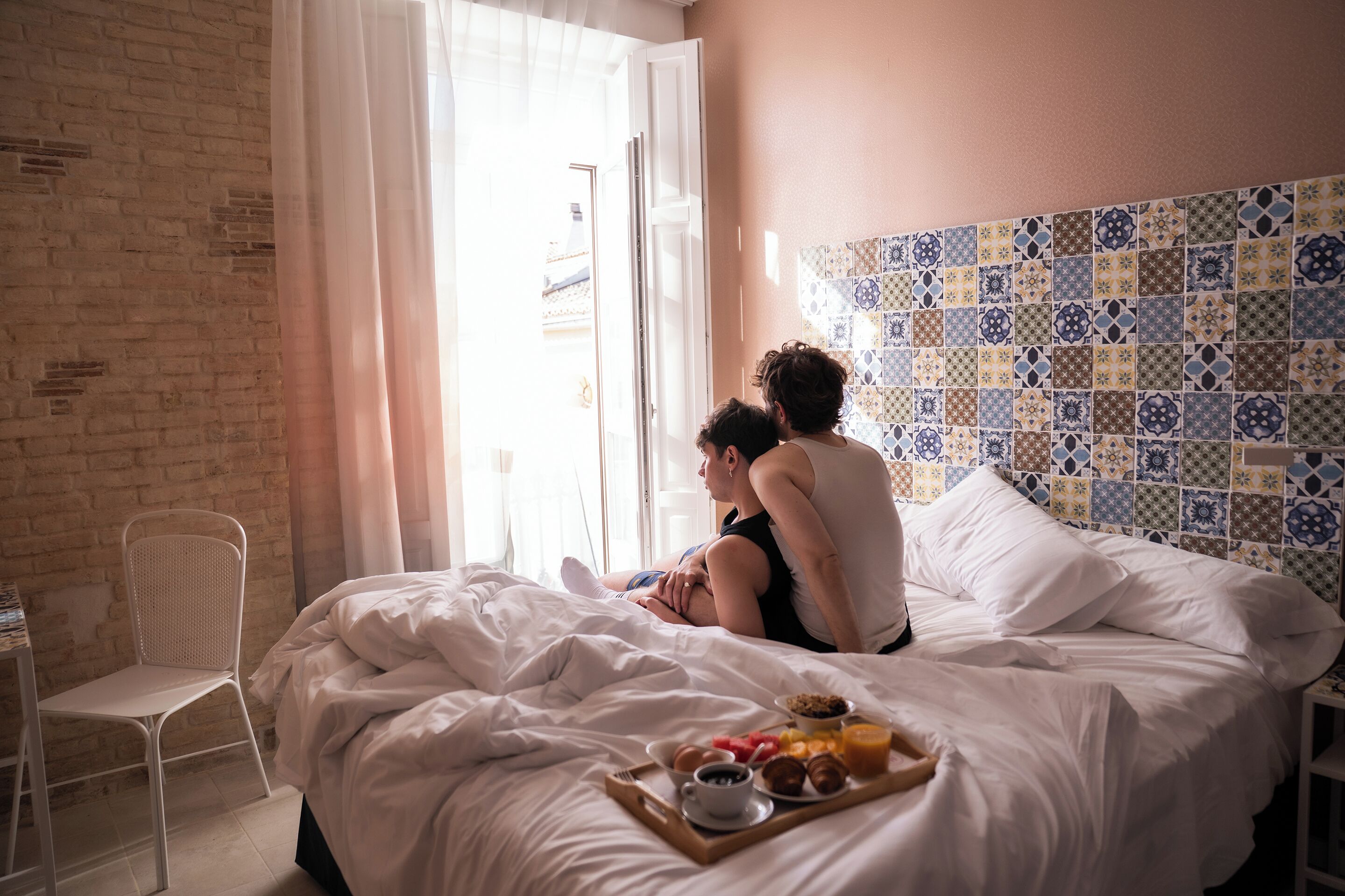 Una cama con una bandeja de desayuno, una silla blanca y una pared de ladrillo.