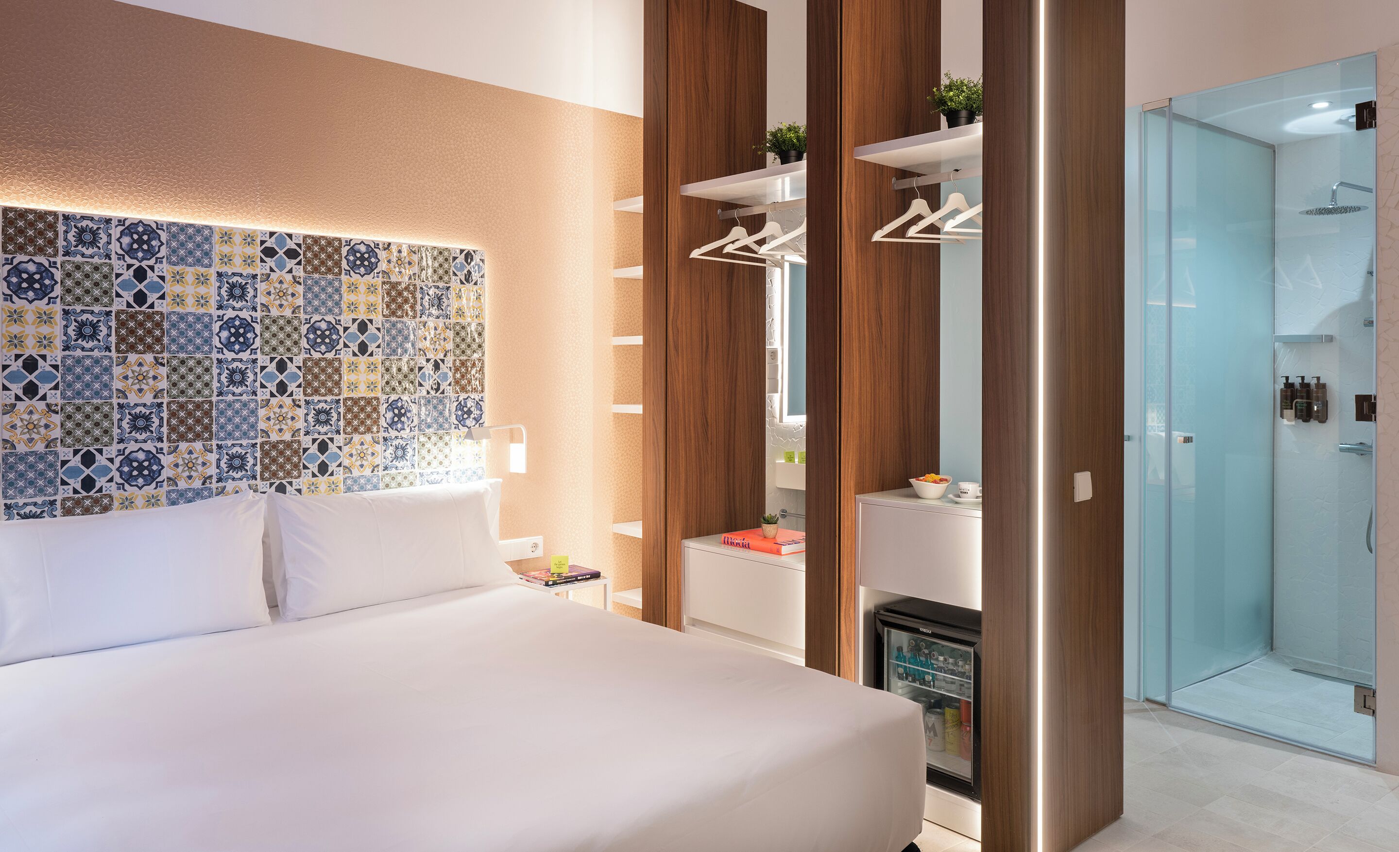 Et moderne hotelværelse med en pænt redt seng, en væg med mønstret fliser, en indbygget garderobe med plads til at hænge tøj, og et brusebad med glasvægge.