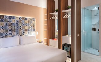 Et moderne hotelværelse med en pænt redt seng, en væg med mønstret fliser, en indbygget garderobe med plads til at hænge tøj, og et brusebad med glasvægge.