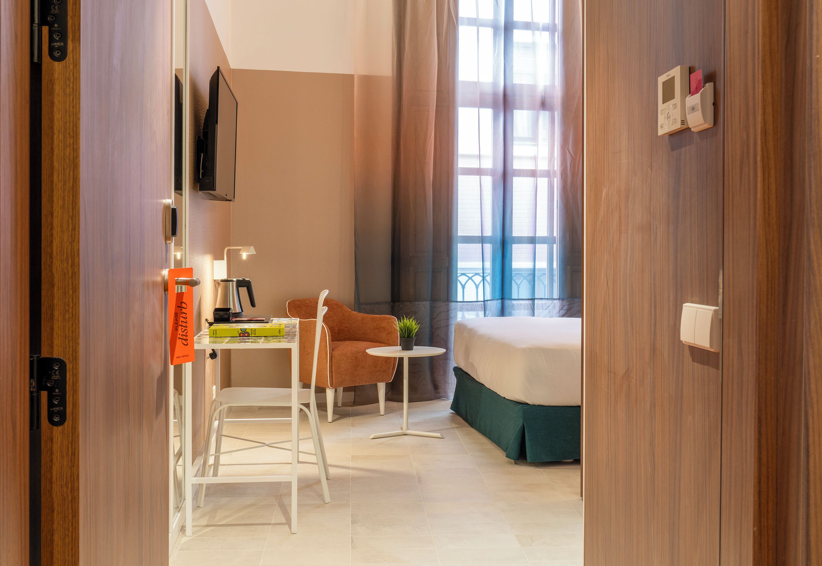 Habitación de hotel con una cama, un escritorio con silla, una mesita con una planta y una ventana con cortinas.