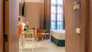 Habitación de hotel con una cama, un escritorio con silla, una mesita con una planta y una ventana con cortinas.