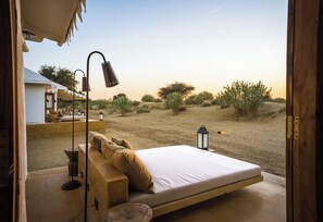 A bedroom with a bed, a nightstand, a lamp, and a view of the desert landscape. - Grand Khalifa Luxury Camp (Jaisalmer)