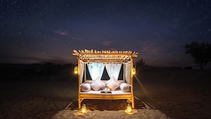 Un lit en bois, agrémenté de draps blancs et d’un dais, éclairé par des lanternes, installé sur une plage de sable fin sous un ciel étoilé.