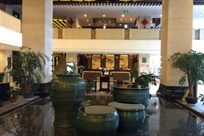 Een ruime lobby met grote groene plantenbakken, een receptiebalie en een hoog plafond met een entresol.