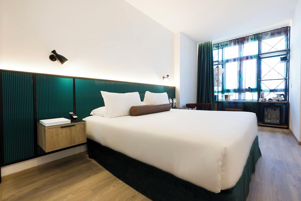 Une chambre d’hôtel avec un grand lit, une table de chevet, une commode, une fenêtre avec des rideaux et une vue sur l’extérieur.