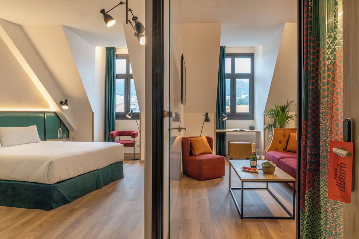 Une chambre d’hôtel moderne avec un lit, un bureau, une chaise et un canapé. Il y a une fenêtre avec des rideaux, une plante et un panneau sur le mur.