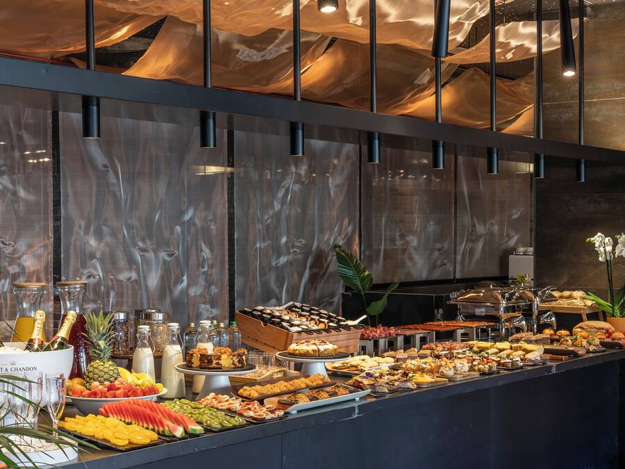 Un bufé con una variedad de alimentos, incluyendo frutas, quesos y carnes, expuestos sobre una barra con una pared y techo de aspecto marmóreo al fondo.