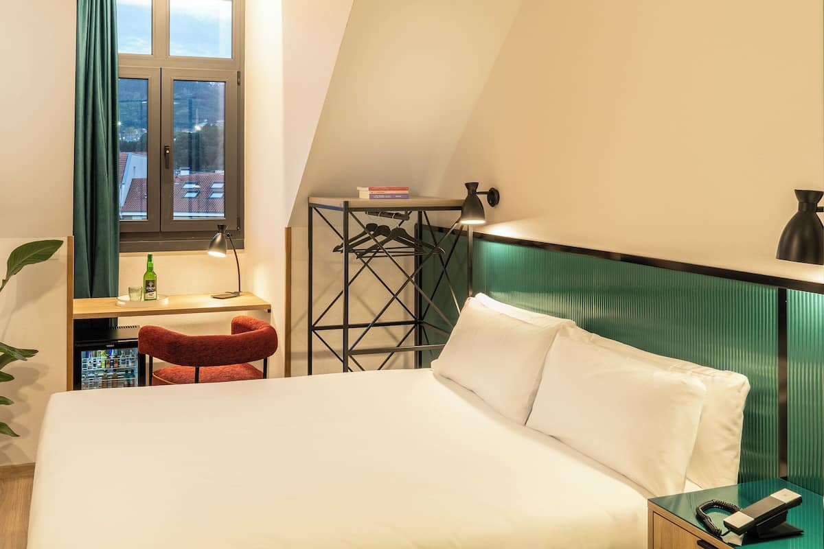 Une chambre d’hôtel compacte comprenant un lit, un petit bureau, une chaise, une plante en pot et une vue sur la ville par la fenêtre.