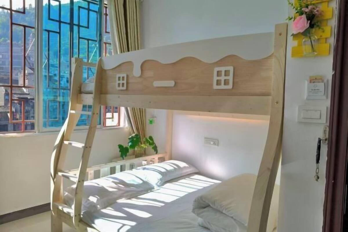 A bunk bed with a wooden headboard, a window with curtains, a bedside plant, and a wall-mounted light switch.